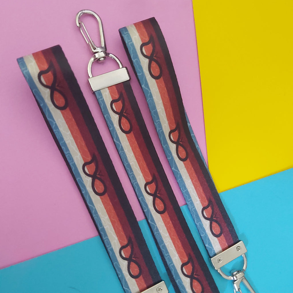 Three fabric strap keychains with silver carabiners, featuring a retro striped pattern of red, blue, white, and brown, displayed on a colourful pink, yellow, and blue background.
