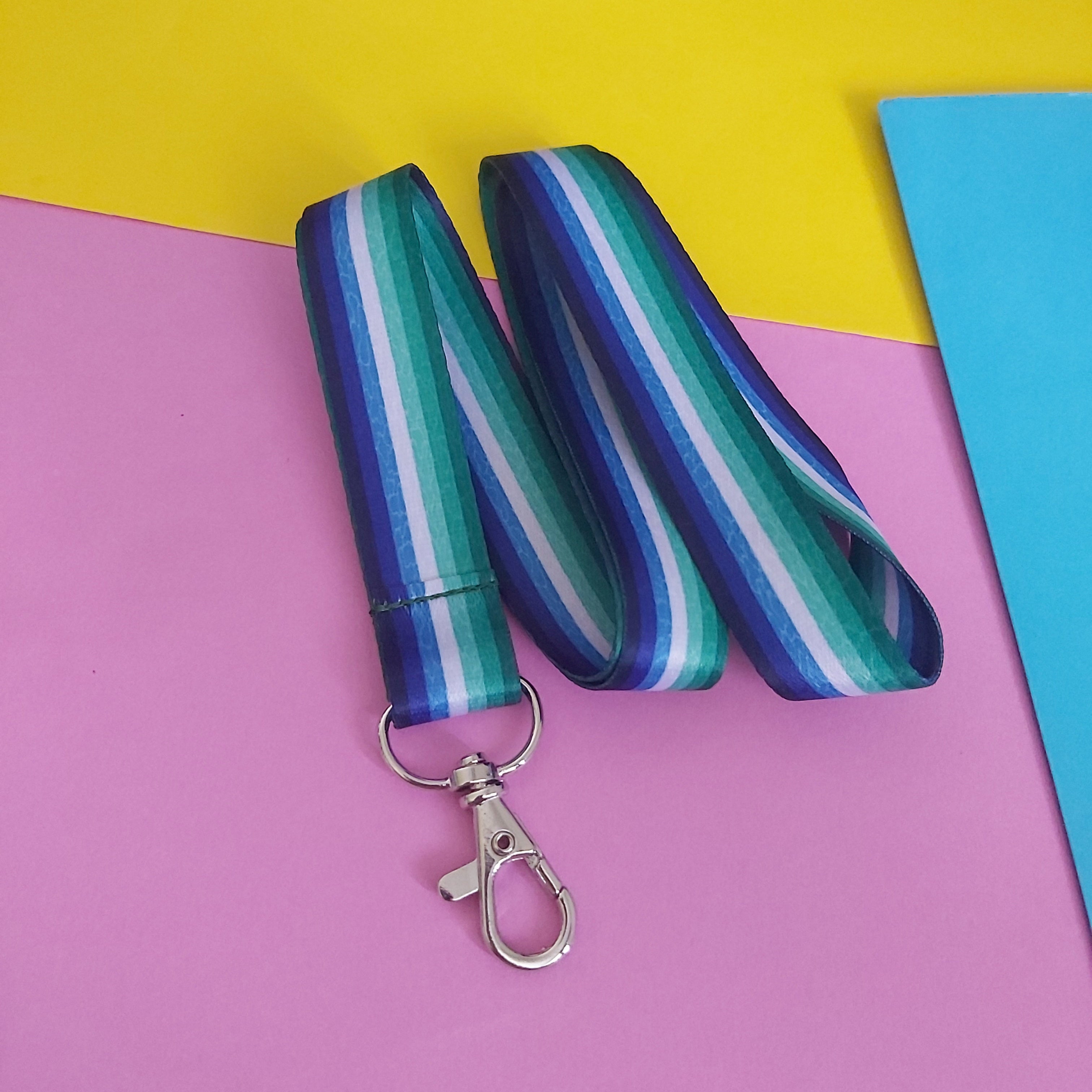 A gay male pride flag striped lanyard with a silver lobster clasp, laid flat on a colorful background of pink, yellow, and blue paper.