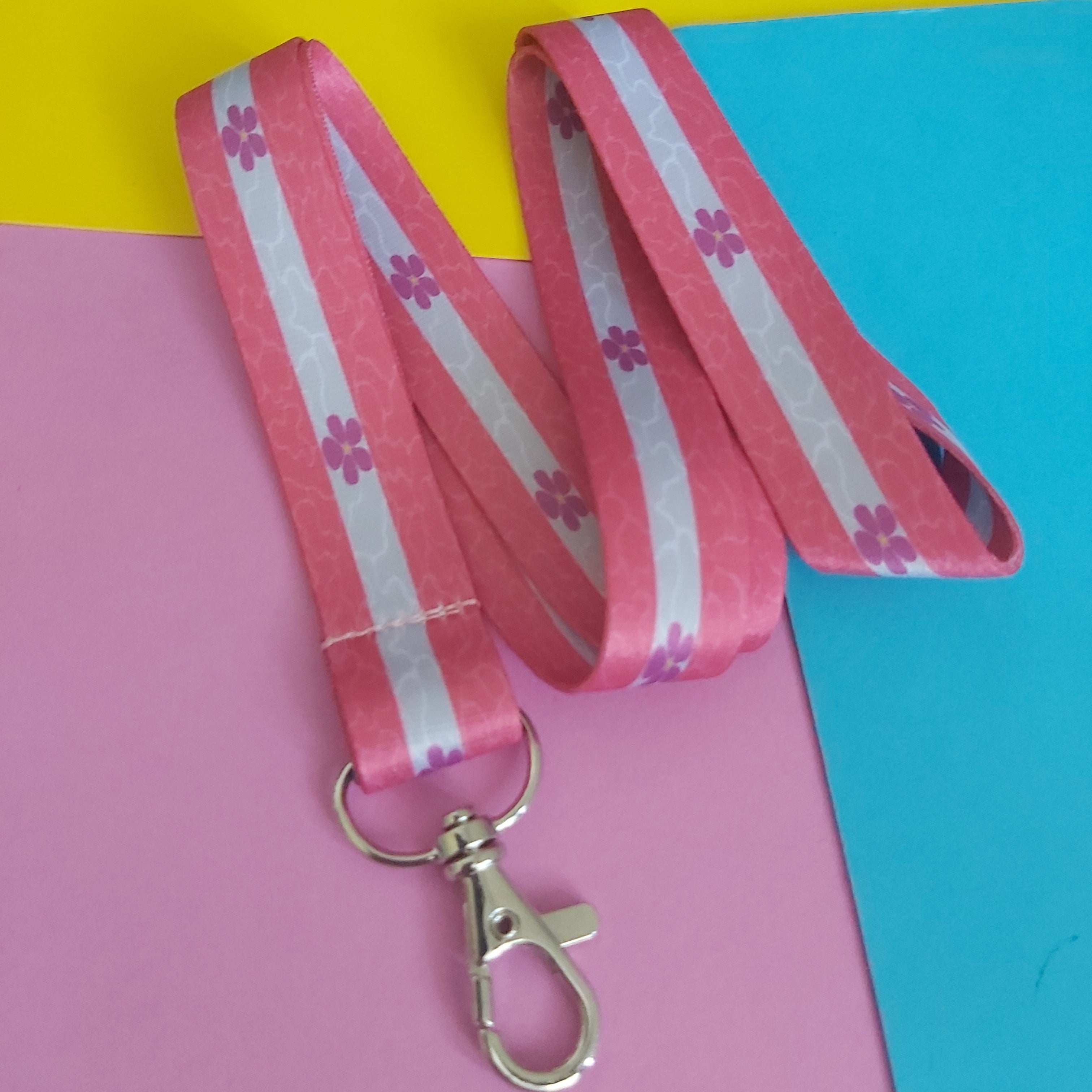A pink and white striped fabric lanyard with small pink flower motifs and a silver metal lobster clasp, laid out on a colourful background of yellow, pink, and blue paper.