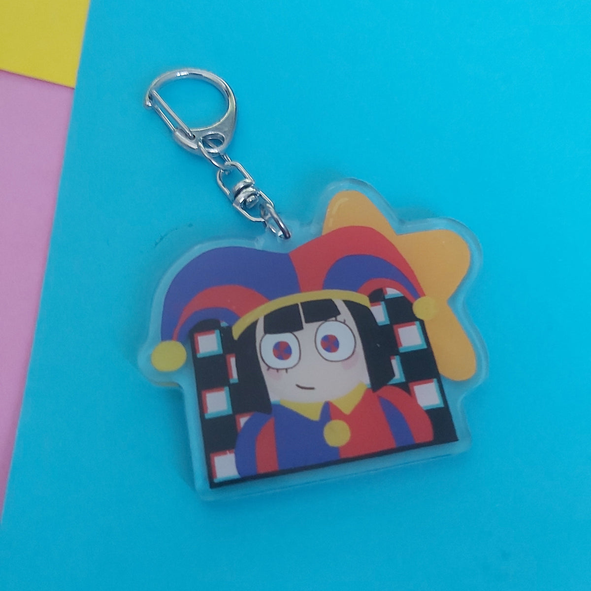 A colorful acrylic keychain featuring a stylized cartoon character with a red, yellow, and blue jester hat, attached to a silver metal clasp, set against a bright blue background with pastel paper accents.