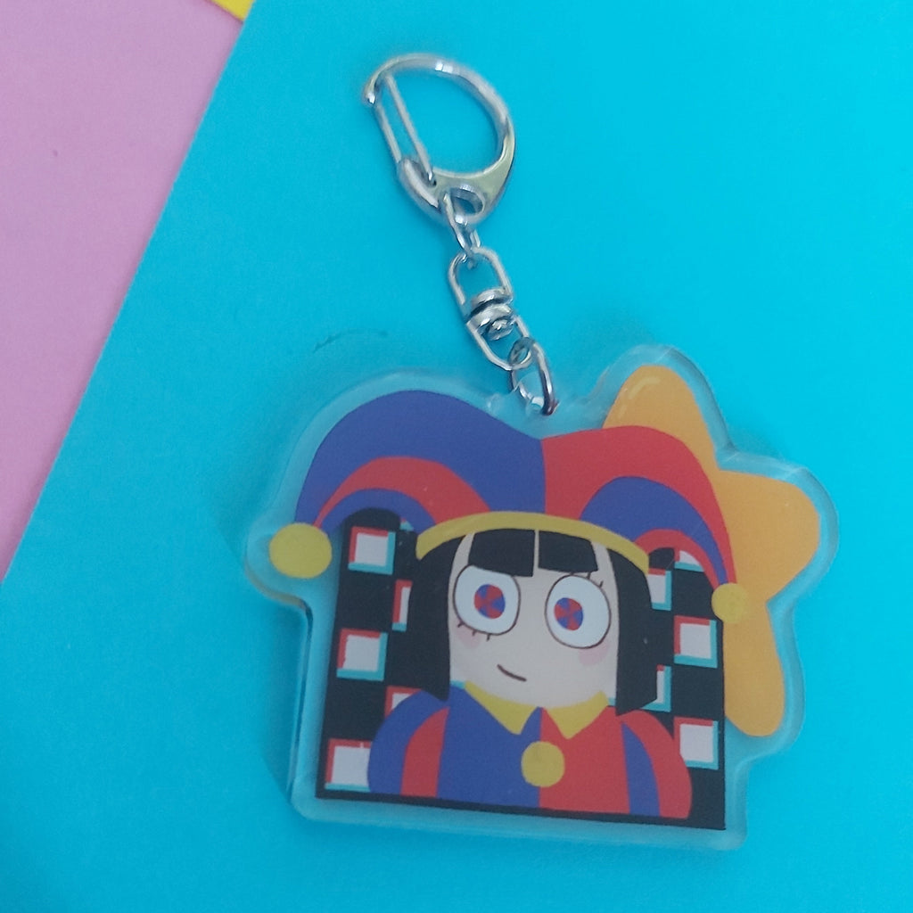 A colorful acrylic keychain featuring a stylized cartoon character with a red, blue, and yellow jester hat, attached to a silver metal clasp, displayed on a bright blue background with pink and yellow corner accents.