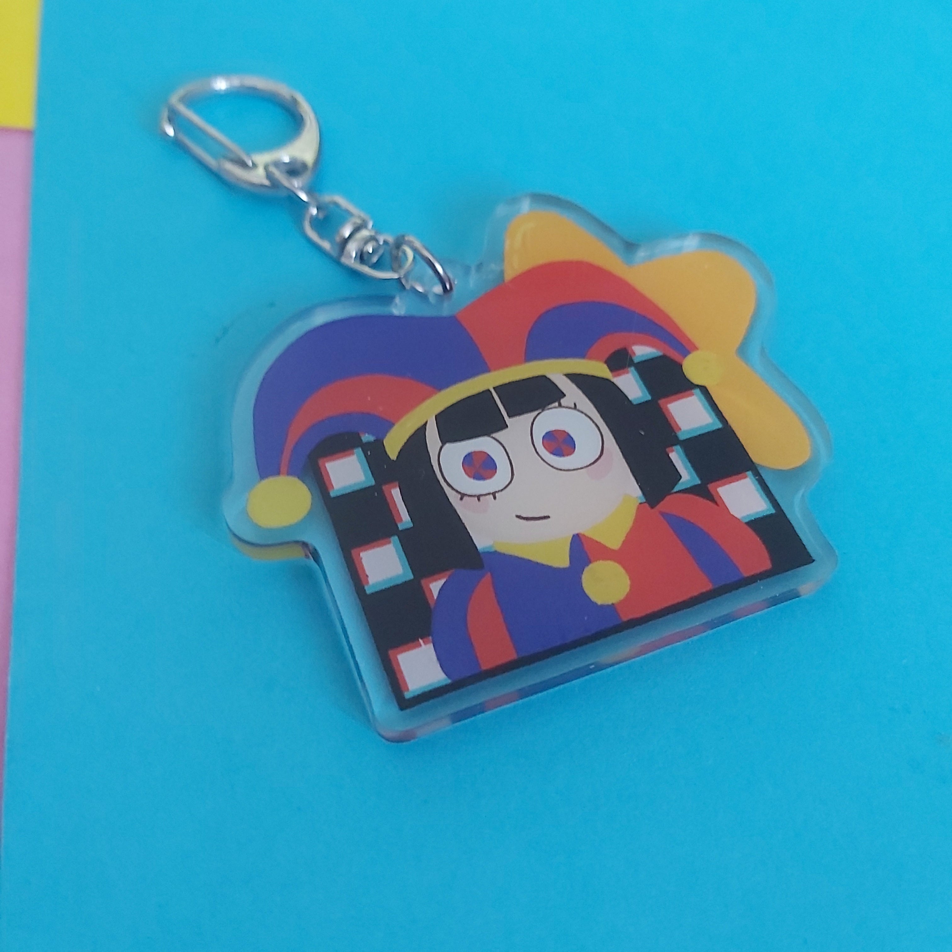 A colorful acrylic keychain featuring an anime-style character with a jester hat, attached to a silver metal clasp, set against a bright blue background.
