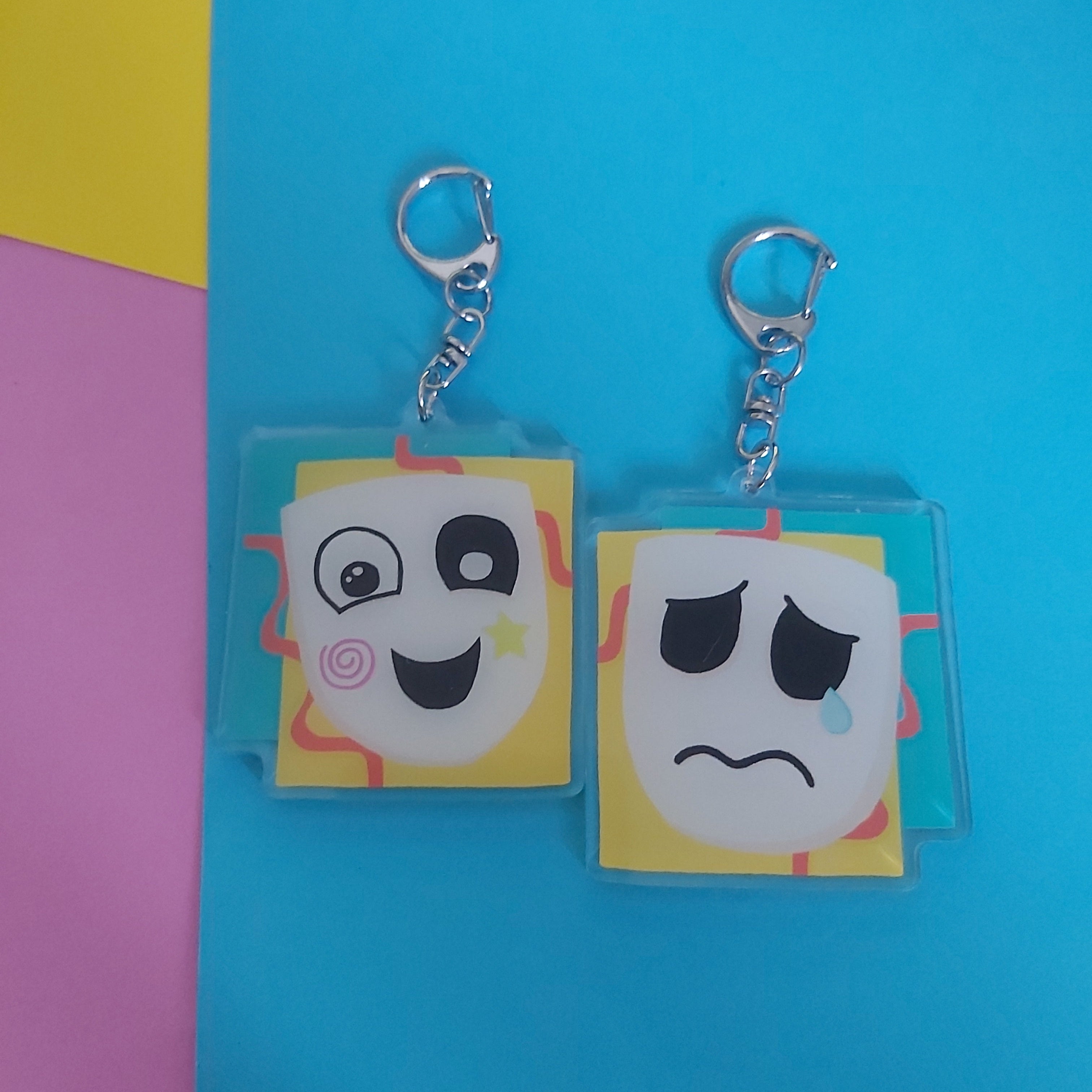 Acrylic keychain with cartoon face charms, showing both sides, one smiling and one sad, on a bright blue background with pink and yellow corner accents.