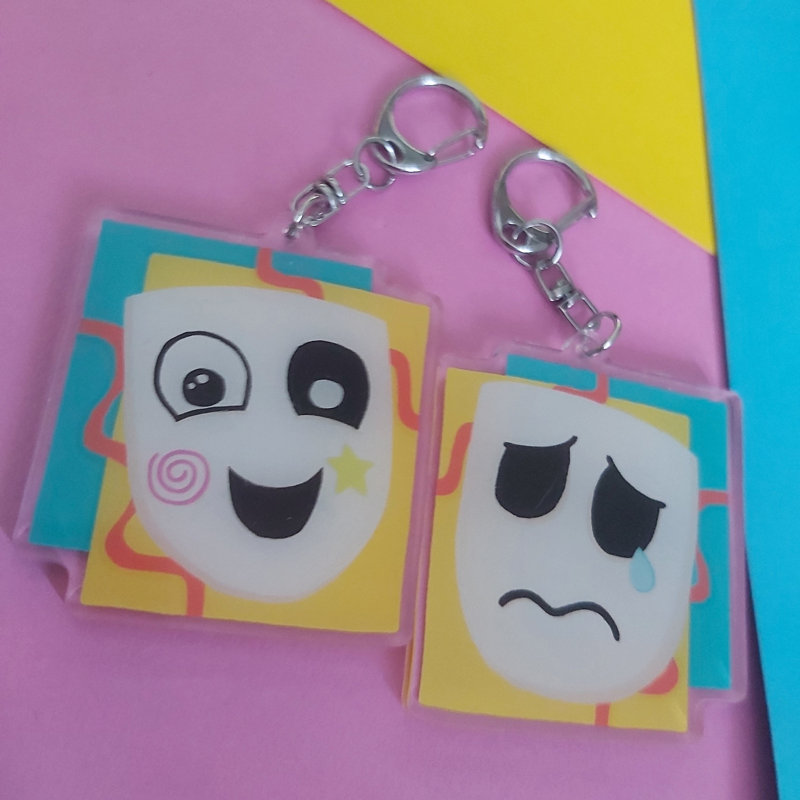 Two sides of acrylic keychains with teary-faced characters on a pink, yellow, and blue background.
