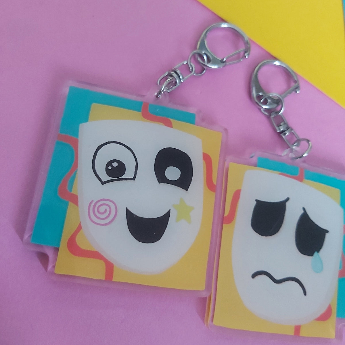 Two acrylic keychains with cartoon mask designs, one smiling and one sad, attached to silver keyrings, displayed on a pink and yellow background.