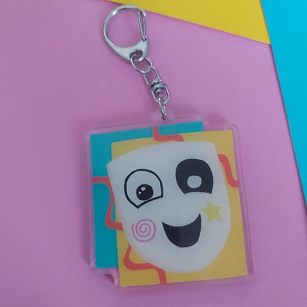A square acrylic keychain featuring a cartoon character with a pink swirl cheek, and yellow star, attached to a silver metal clasp. The keychain is photographed on a background of pink, yellow, and blue paper.