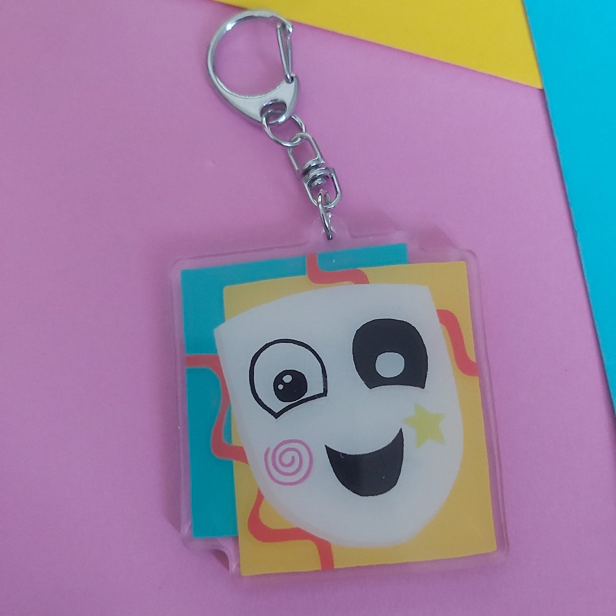 A square acrylic keychain featuring a cartoon character with a pink swirl cheek, and yellow star, attached to a silver metal clasp. The keychain is photographed on a background of pink, yellow, and blue paper.