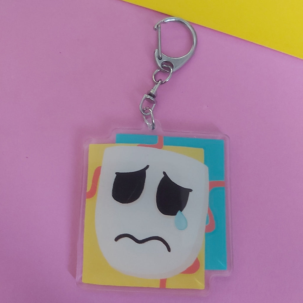 A square acrylic keychain charm featuring a sad theatre mask face with a blue tear, attached to a silver metal lobster clasp, set against a pink and yellow background.