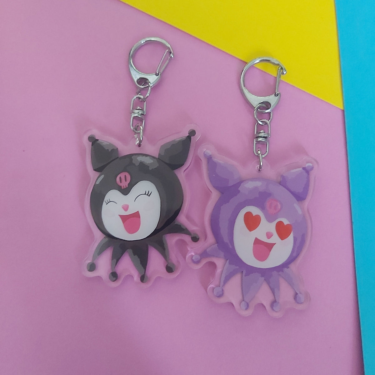 Cute cartoon keychains with silver metal clips, one side black and one purple, displayed on a pink background with yellow and blue corner accents.