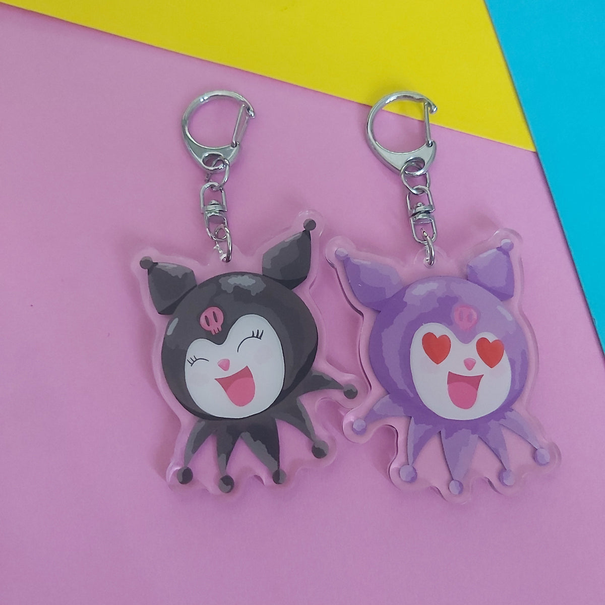 Two cute cartoon character acrylic keychains on a colourful pastel background. One keychain is black with a white face and pink details, and the other is light purple with a white face and pink heart eyes.