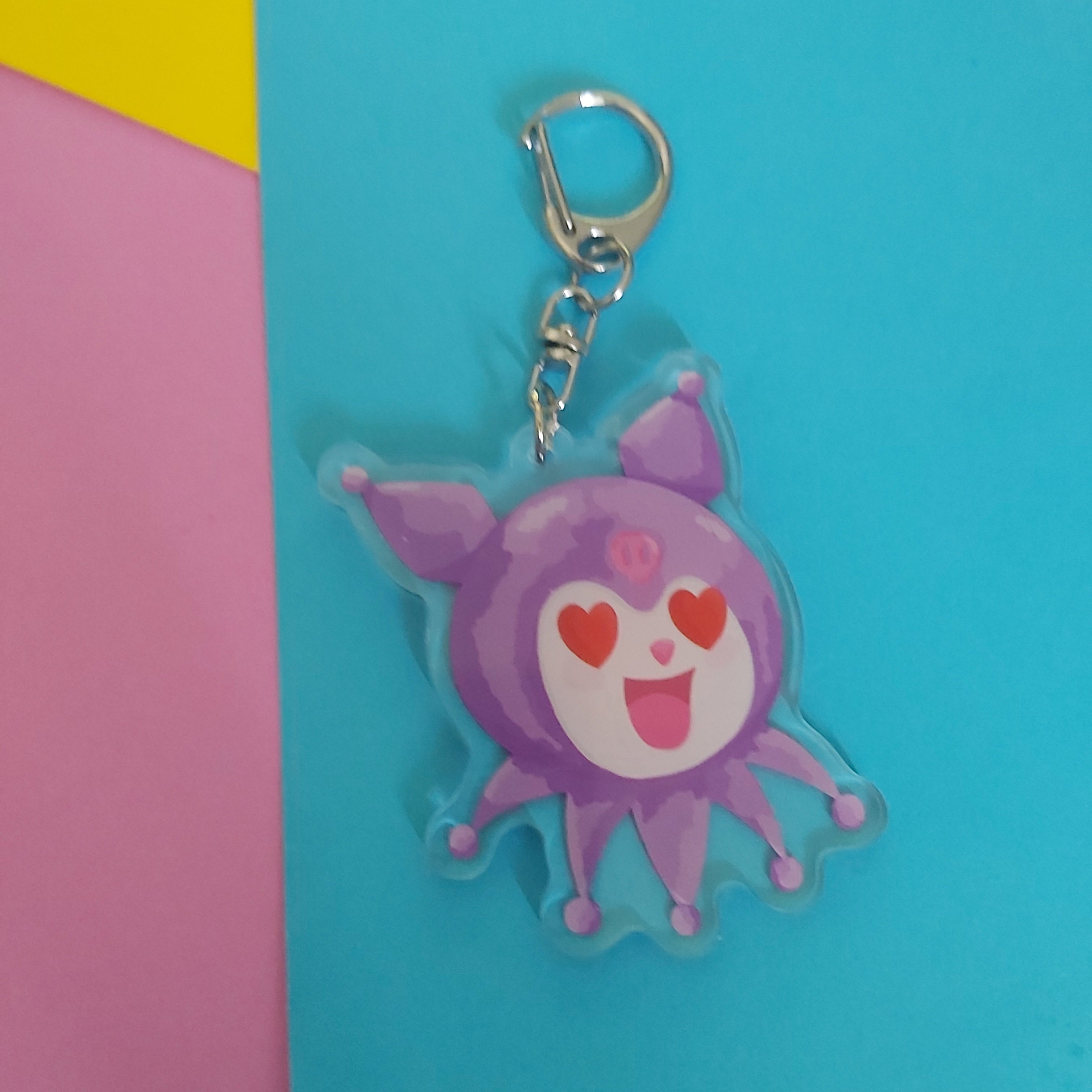 A purple cartoon character keychain charm with red heart eyes and a silver metal clasp, shown against a bright blue background with pink and yellow color blocks on the left.