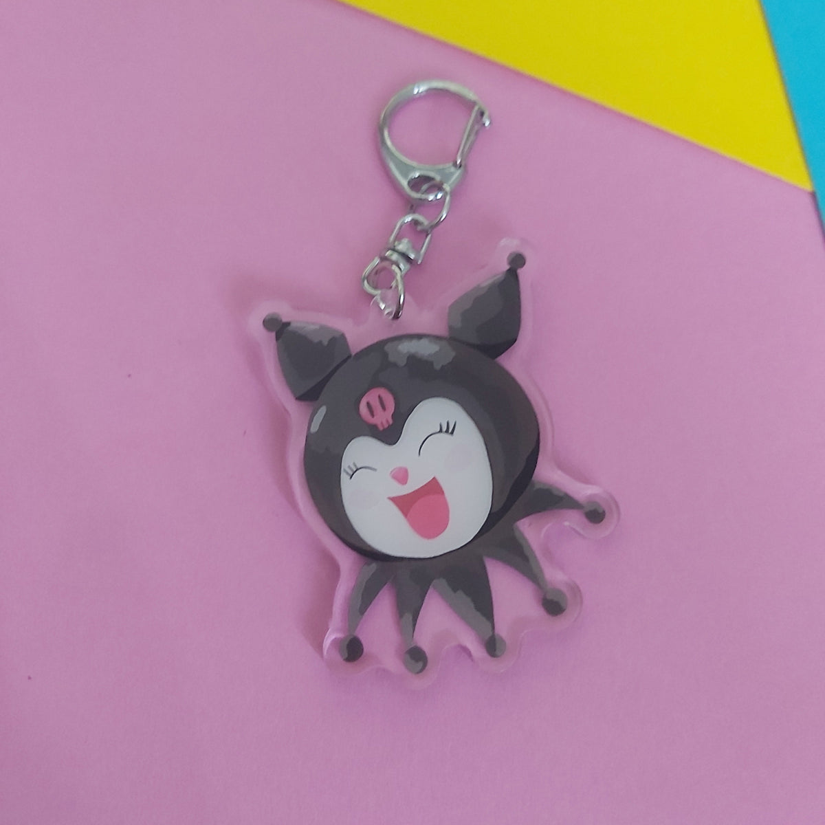 A cartoon character keychain charm with a silver clasp, featuring a black and white character with pink details, displayed on a pink background with yellow and blue corner accents.