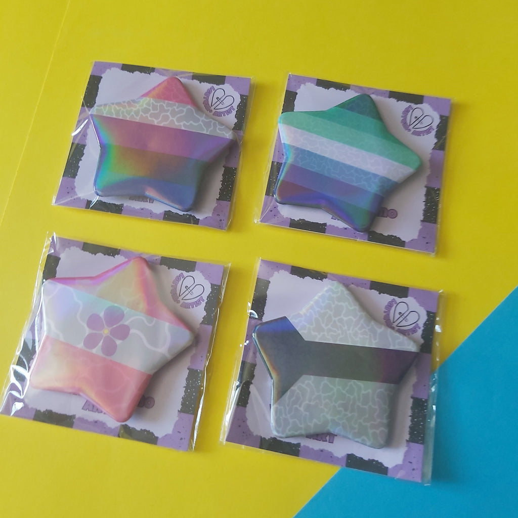 Four star-shaped pinback buttons with the sapphic, genderfluid, gay male and demisexual pride flags, each in its own clear plastic sleeve on a bright yellow and blue background.