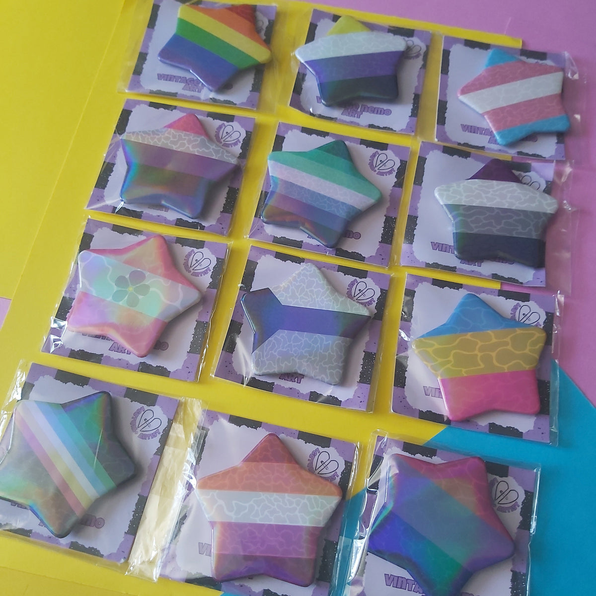 Twelve star-shaped rainbow pins, each individually wrapped in clear plastic and attached to a printed backing card, arranged on a bright yellow and blue surface with a pink corner accent.