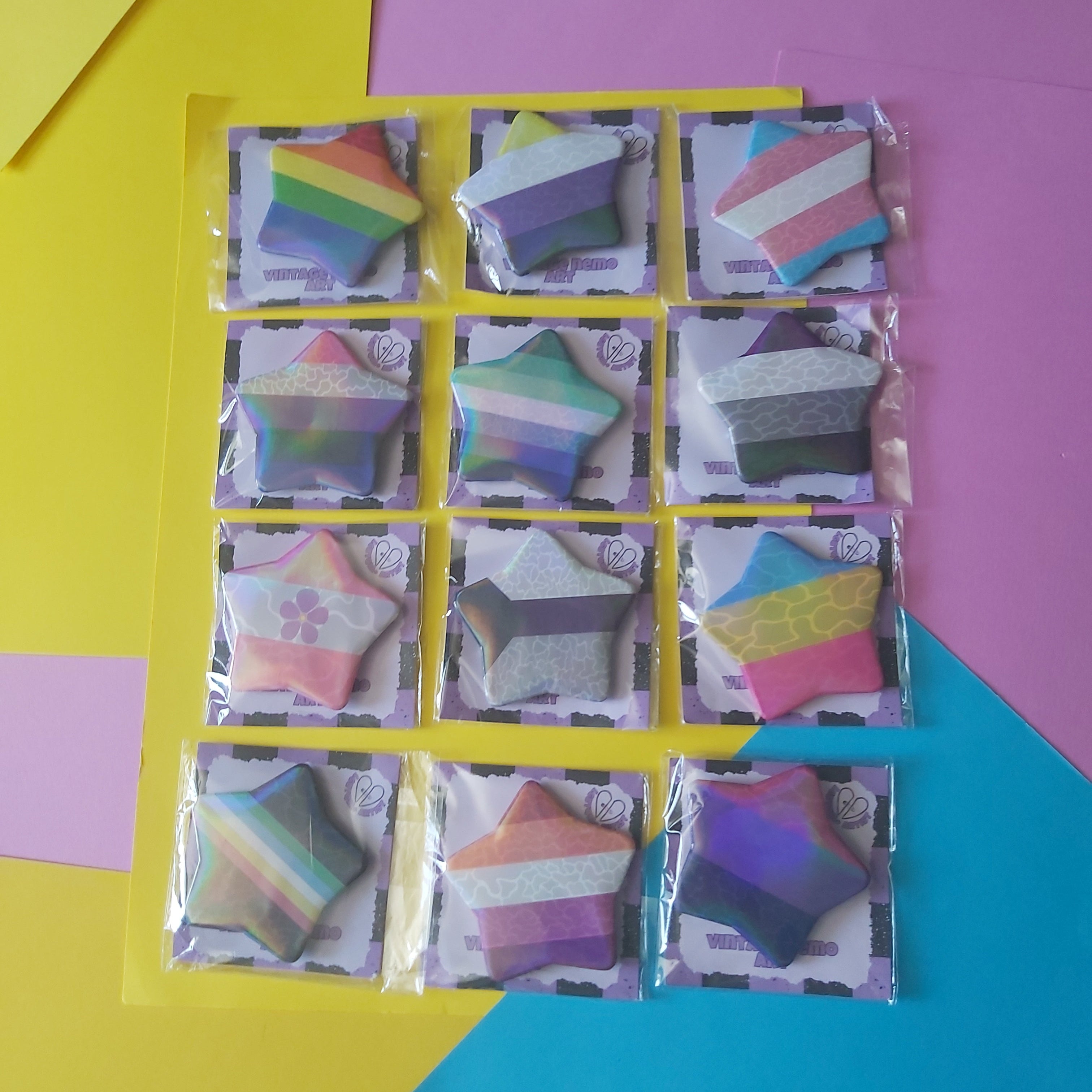 A set of 12 star-shaped badges in various pride flags packaged in clear plastic sleeves and arranged on a yellow card over a colourful geometric background of yellow, pink, and blue paper.