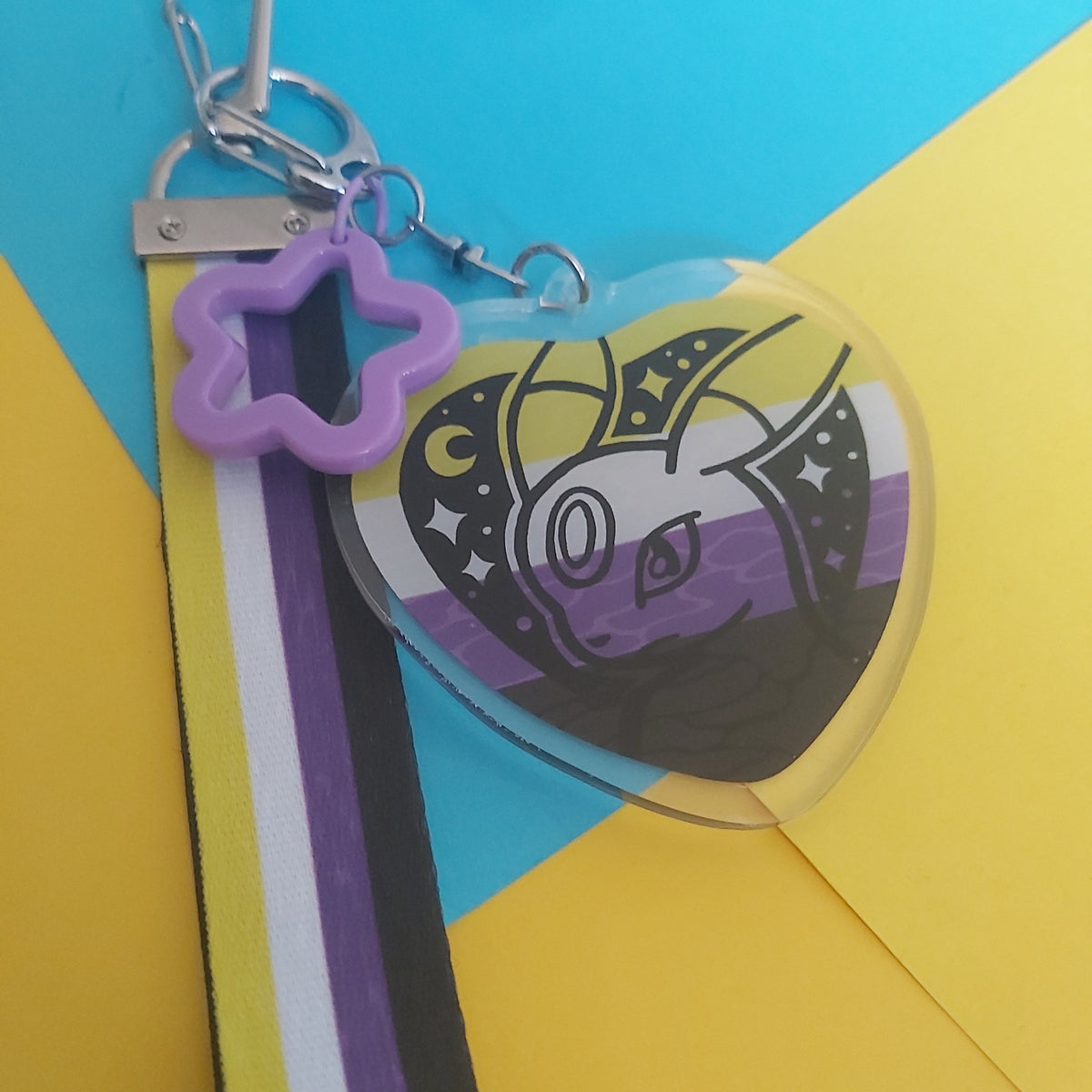 A heart-shaped acrylic keychain featuring a Pokémon with a purple, black, and yellow striped design, attached to a purple star charm and a matching striped ribbon on a yellow and blue background.