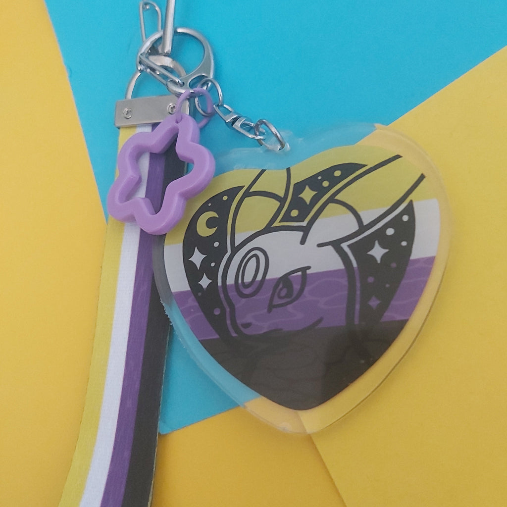 A heart-shaped acrylic keychain featuring a stylised Pokémon illustration in a purple, black, white, and yellow design, attached to a silver clasp with a purple star charm and a striped ribbon.