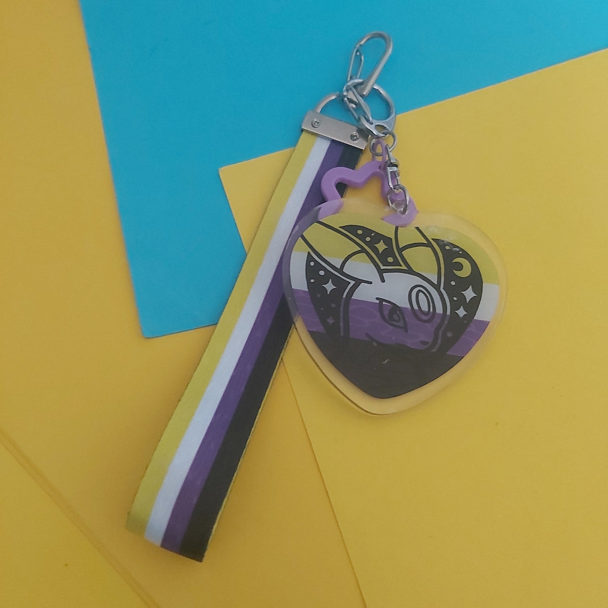 A heart-shaped acrylic keychain charm featuring a stylized purple and black graphic, attached to a striped fabric wristlet strap with yellow, white, purple, and black bands, displayed on a yellow and blue background.