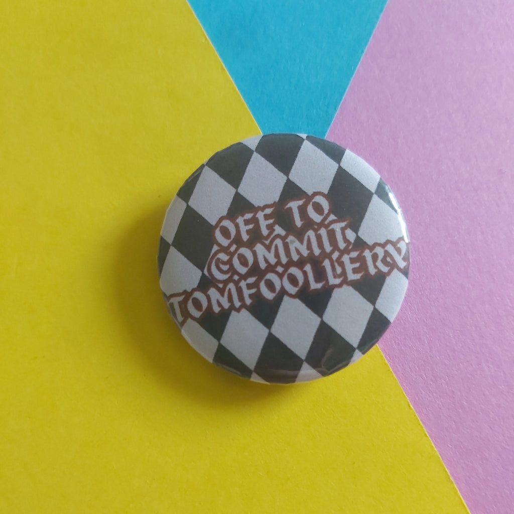 A round pin badge with a black and white checkered pattern and the text 'OFF TO COMMIT TOMFOOLERY' in red, placed on a diagonally split yellow, blue, and pink background.