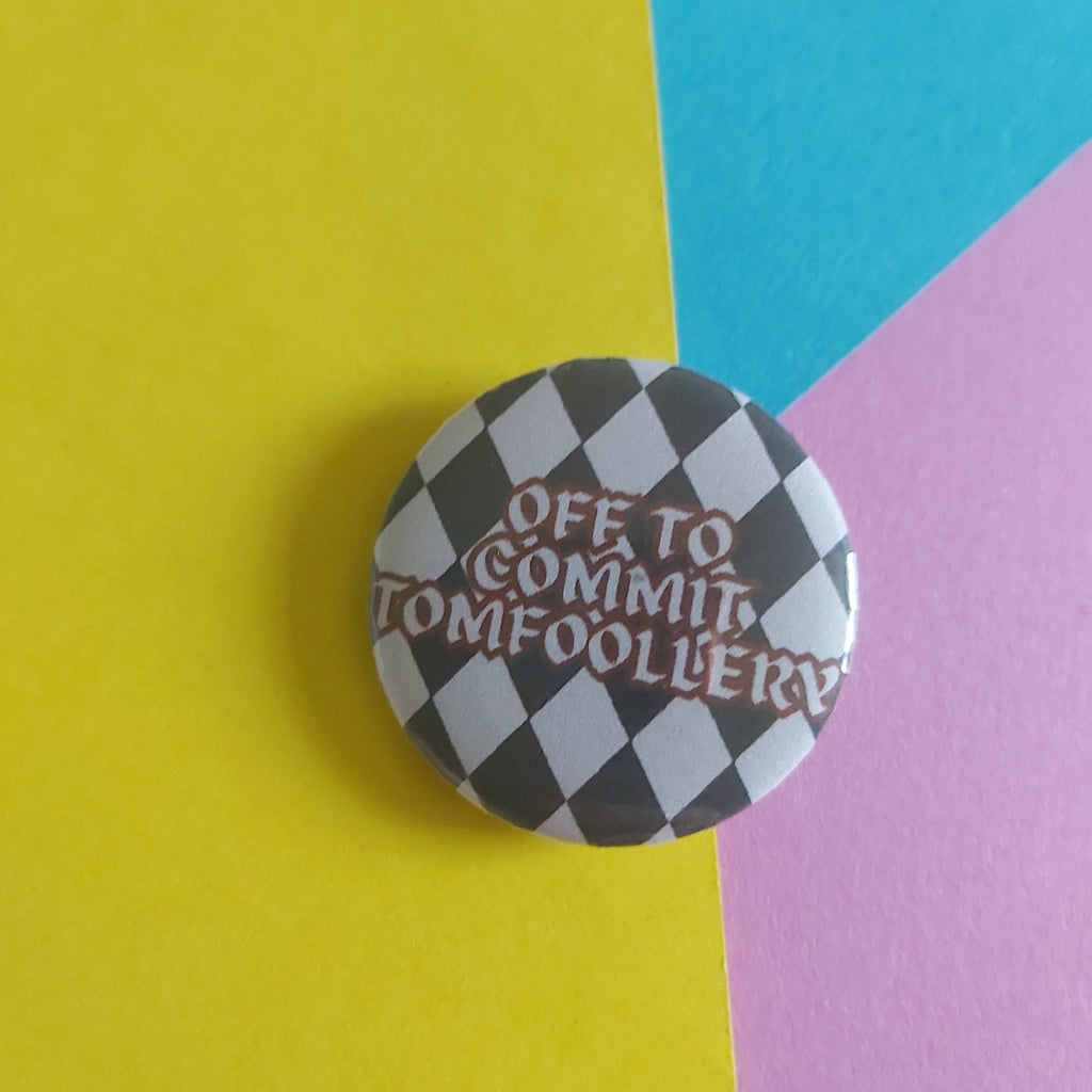 A round pin badge with a black and white checkered pattern and the text 'OFF TO COMMIT TOMFOOLERY' in red, placed on a diagonally split yellow, blue, and pink background.
