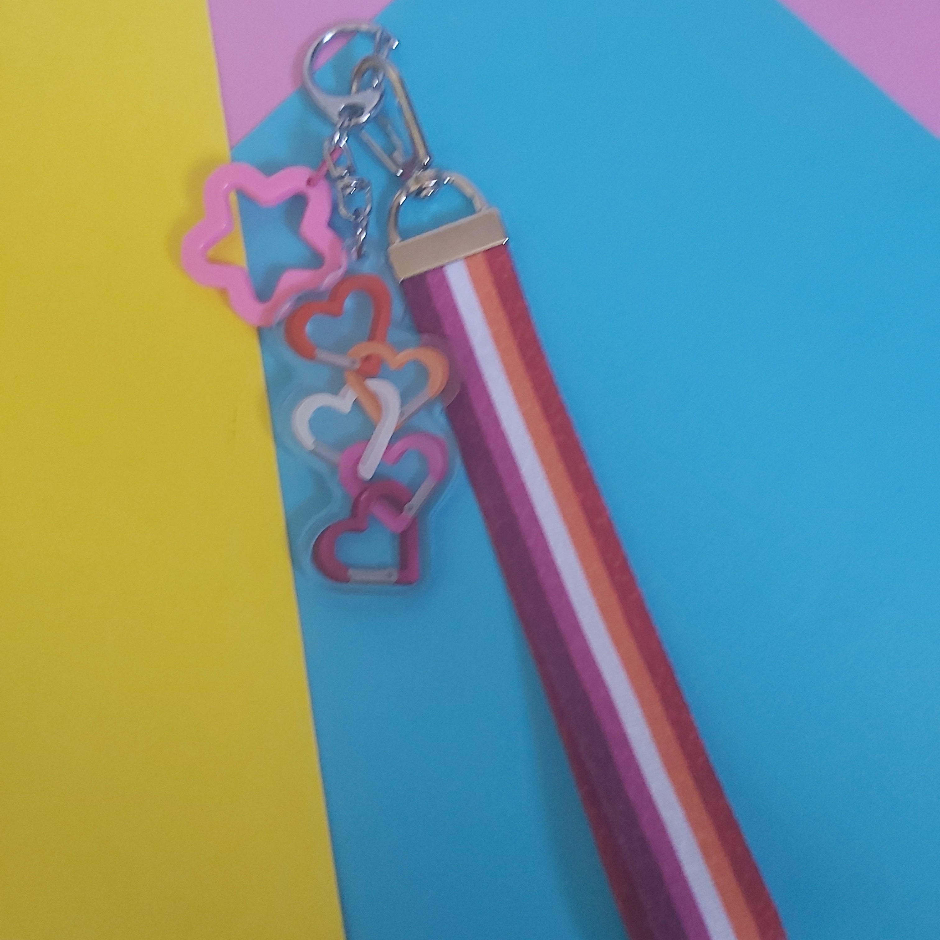 A striped keychain lanyard with a silver clasp and dangling acrylic charm in pink, orange, and white, displayed on a bright blue sheet of paper with a yellow sheet in the background.