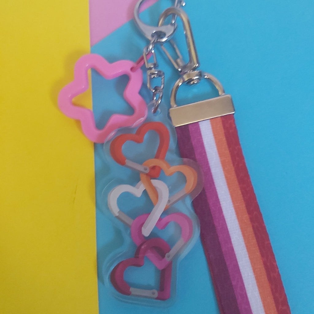 A colourful keychain with a silver clasp, featuring a pink star-shaped charm and a charm with interlocking heart carabiners in pink, orange, and white. It is attached to a striped fabric strap in pink, orange, and white, set against a diagonally split yellow, pink, and blue background.