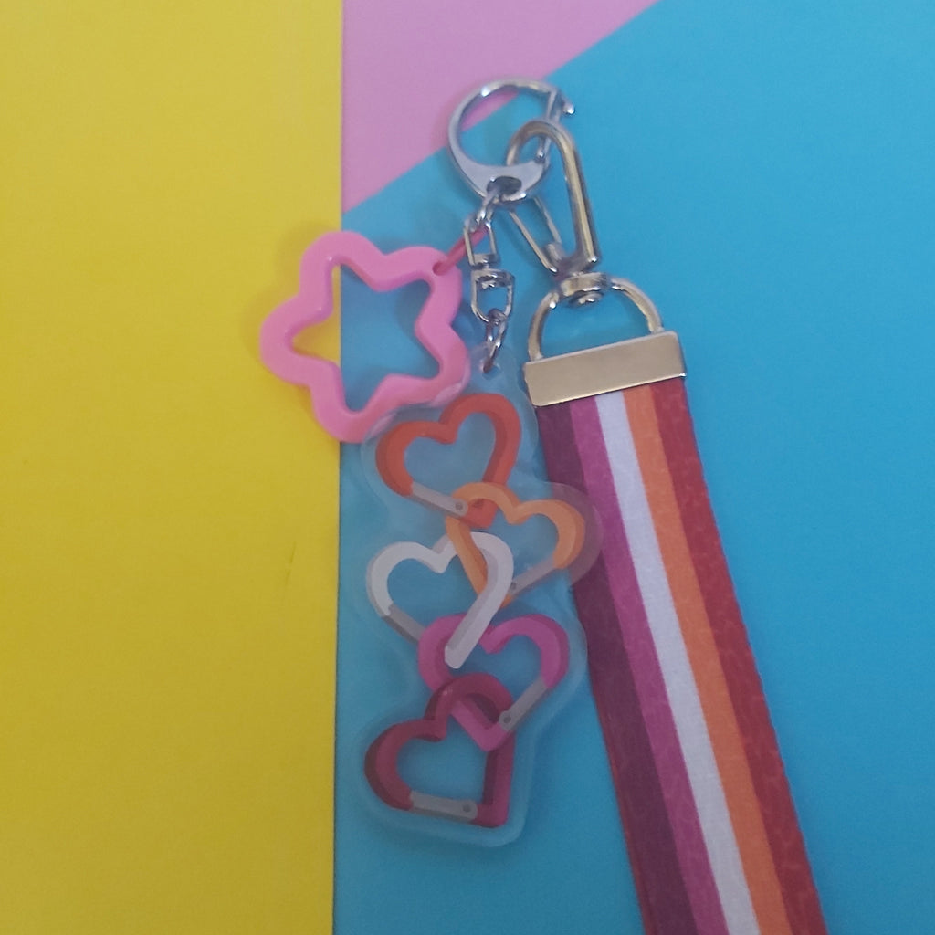 A colourful keychain with a pink star-shaped charm, a stack of interlocking heart charms in pink, orange, and white, and a striped wristlet strap in pink, orange, and white, displayed on a background of yellow and blue paper.