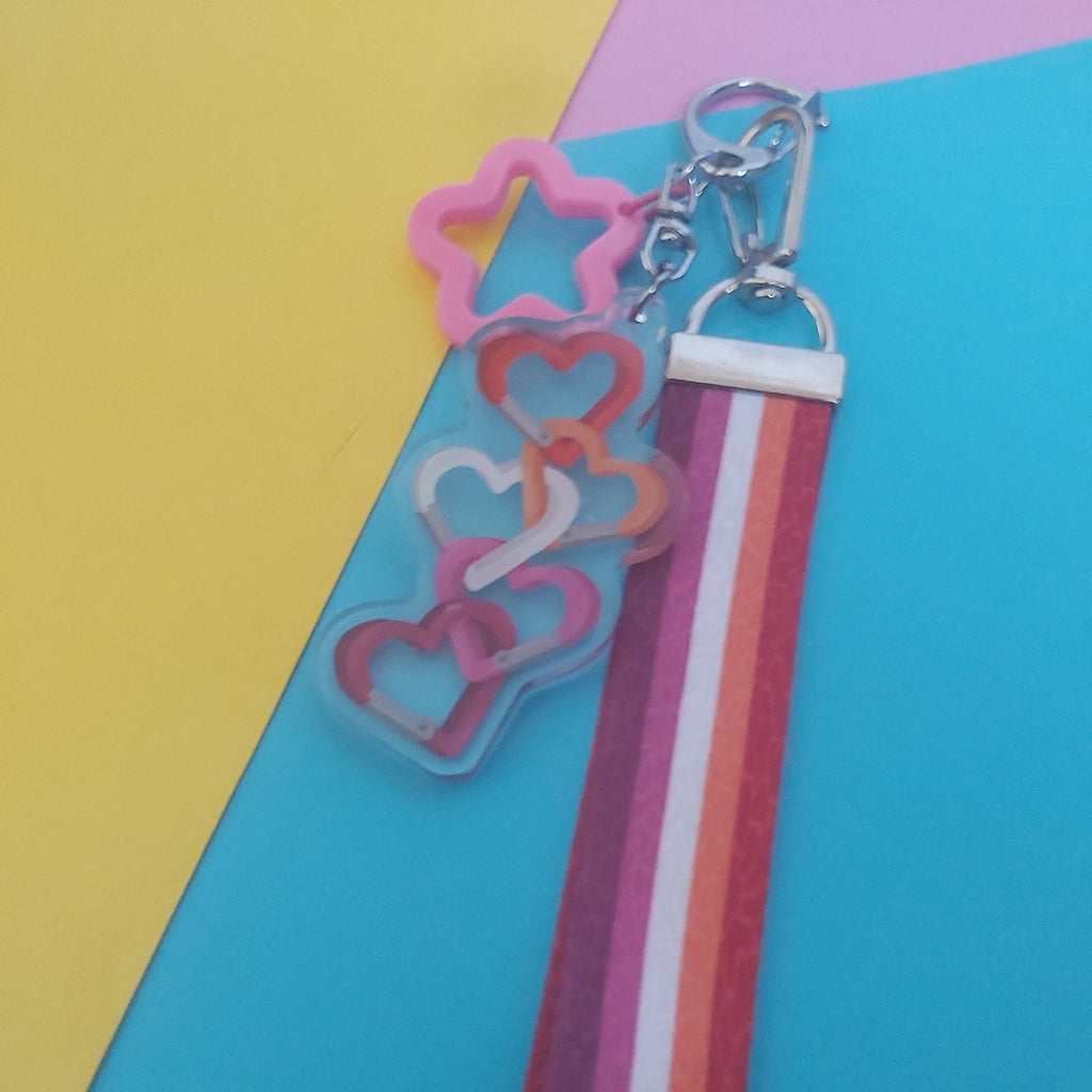 A wristlet strap keychain with a colourful striped fabric strap and interlocking heart-shaped acrylic charms in pink, orange, and red, displayed on a background of yellow, blue, and pink paper.