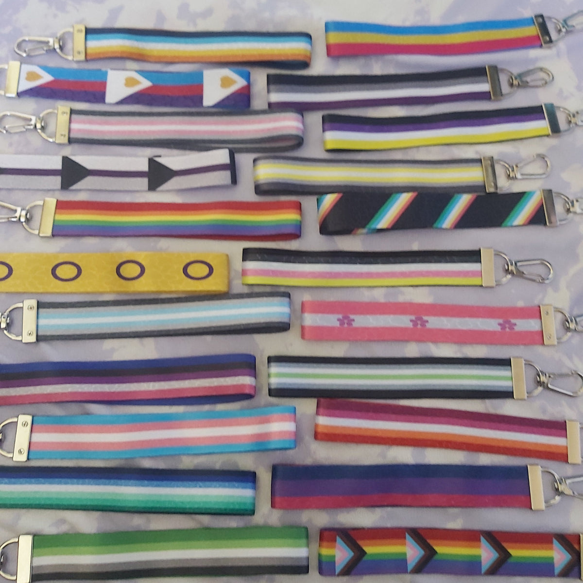 A collection of colourful fabric keychain wristlets with silver metal lobster clasps, arranged in rows on a light purple and white marbled surface. The lanyard features various pride flags