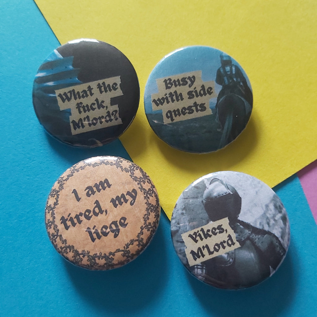 Four round pin-back buttons with humorous text designs arranged on a colourful background of blue and yellow paper. The designs feature medieval-themed graphics and text, including 'What the fuck, M'lord?', 'Busy with side quests', 'I am tired, my liege', and 'Yikes M'Lord'.