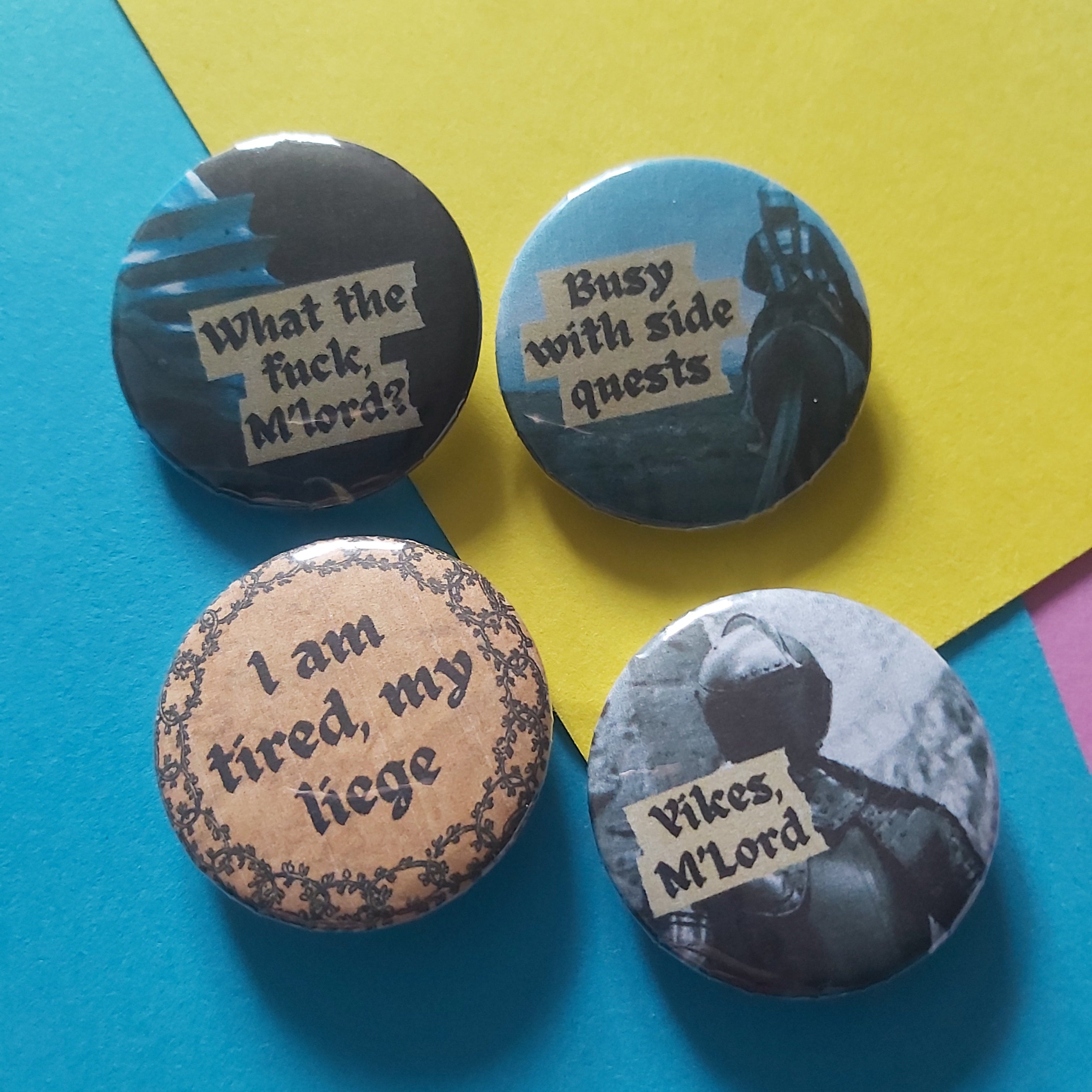 Four round pin-back buttons with humorous text designs arranged on a colourful background of blue and yellow paper. The designs feature medieval-themed graphics and text, including 'What the fuck, M'lord?', 'Busy with side quests', 'I am tired, my liege', and 'Yikes M'Lord'.