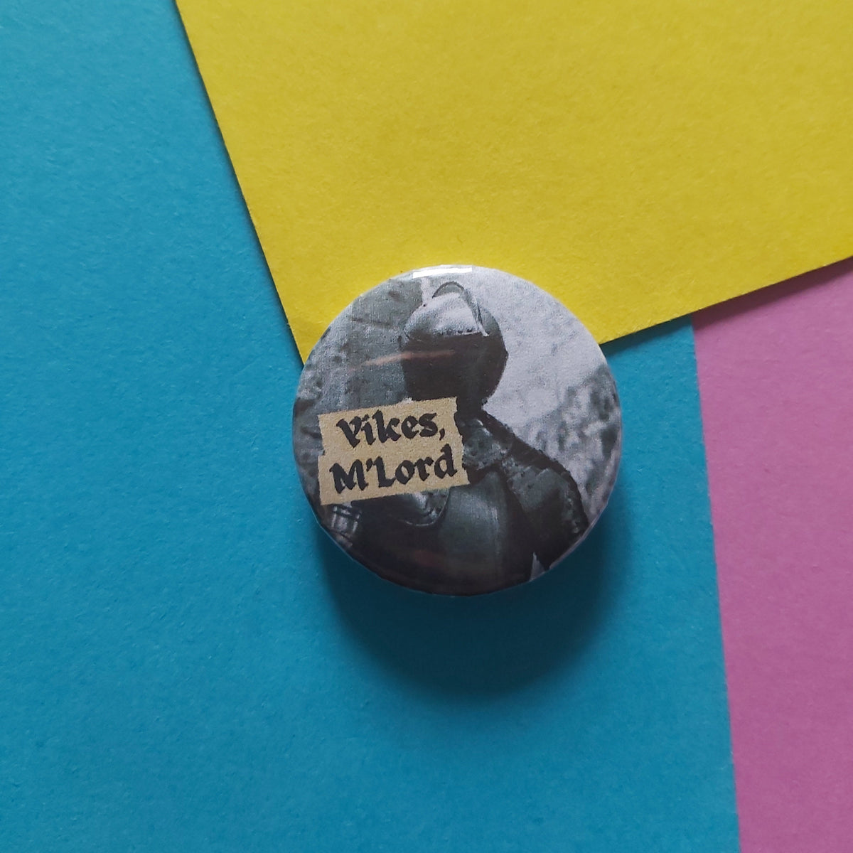 A round pin-back button badge featuring a black-and-white historical illustration of a Viking with a paper label reading 'Yikes, M'Lord', placed on a colourful background of blue, yellow, and pink paper.