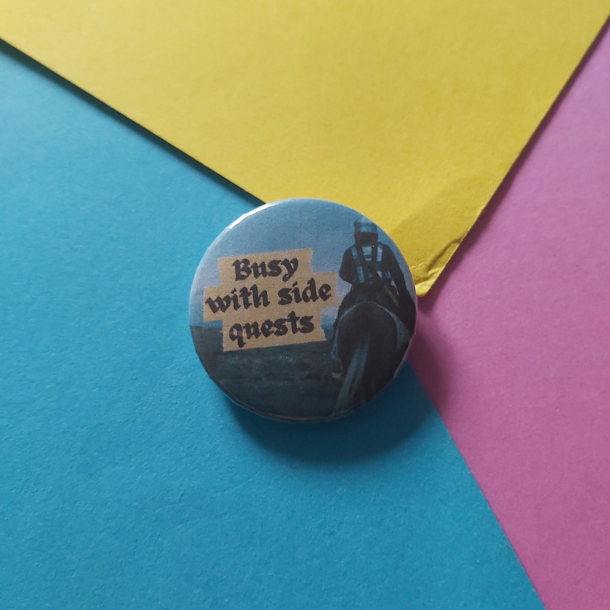 A round pinback button badge featuring a dark illustration of a figure on horseback and the text 'Busy with side quests' in beige lettering, placed on a colorful background of blue, yellow, and pink paper.
