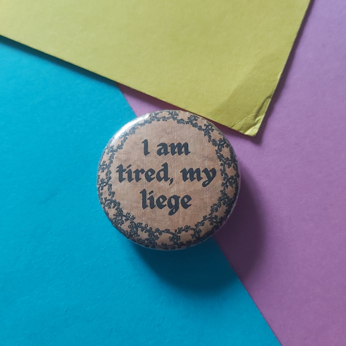 A round pinback button with the text 'I am tired, my liege' in black script on a beige background with a dark floral border, placed on a colorful background of blue, purple, and yellow paper.