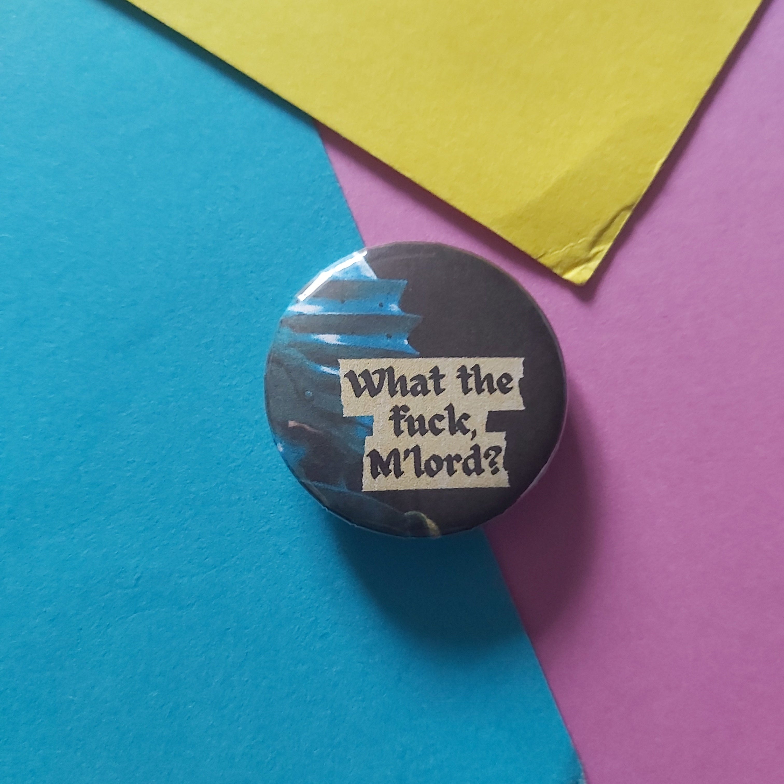 A round pinback button badge with a dark background and the text 'What the fuck, M'lords?' in white, placed on a colorful background of blue, yellow, and pink paper.