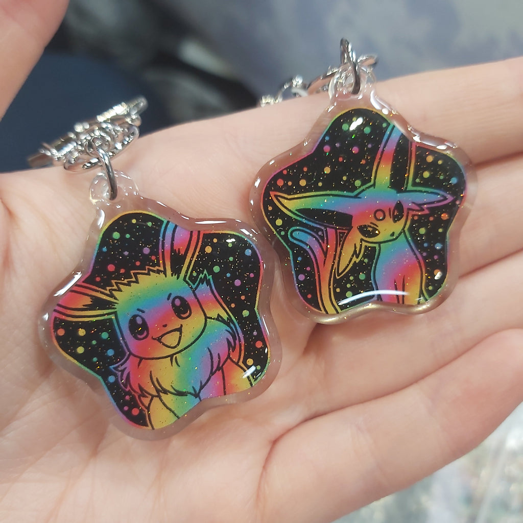 Two star-shaped keychains featuring colorful holographic rainbow designs of a cute creature on a black starry background, held in a person's hand.