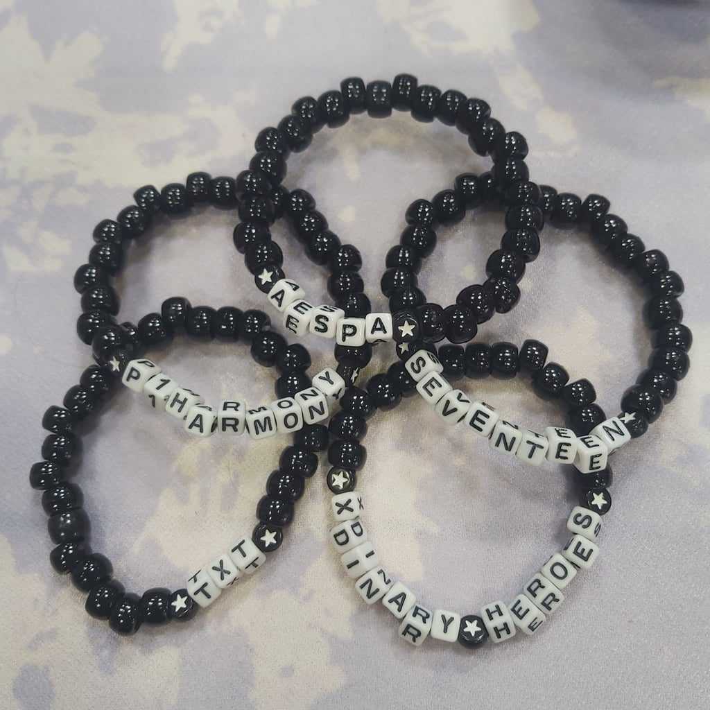 Five black beaded stretch bracelets with white letter beads spelling out K-pop group names, arranged on a light purple and white patterned surface.