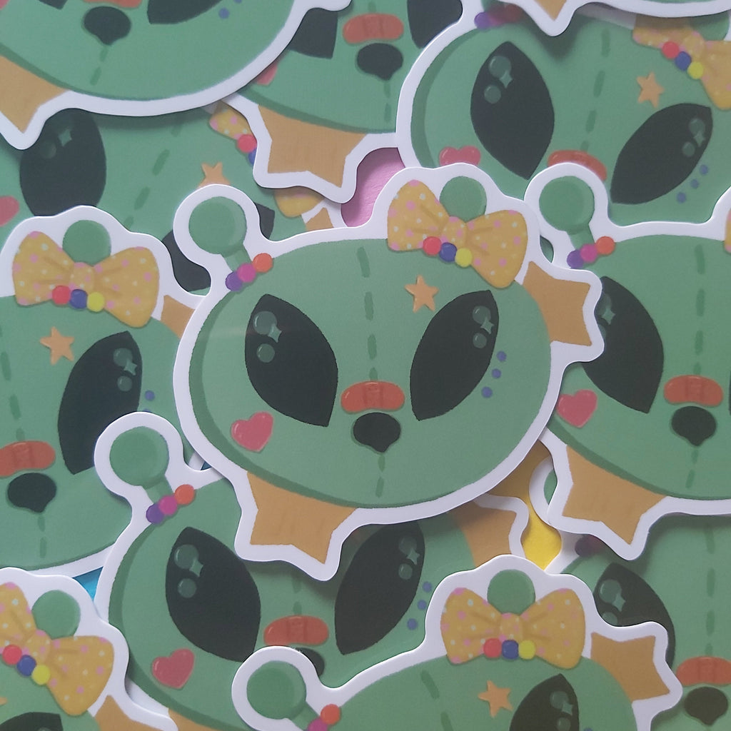 A pile of cartoon alien stickers with large black eyes, a yellow polka-dot hair bow, and small colourful accents, photographed close up.
