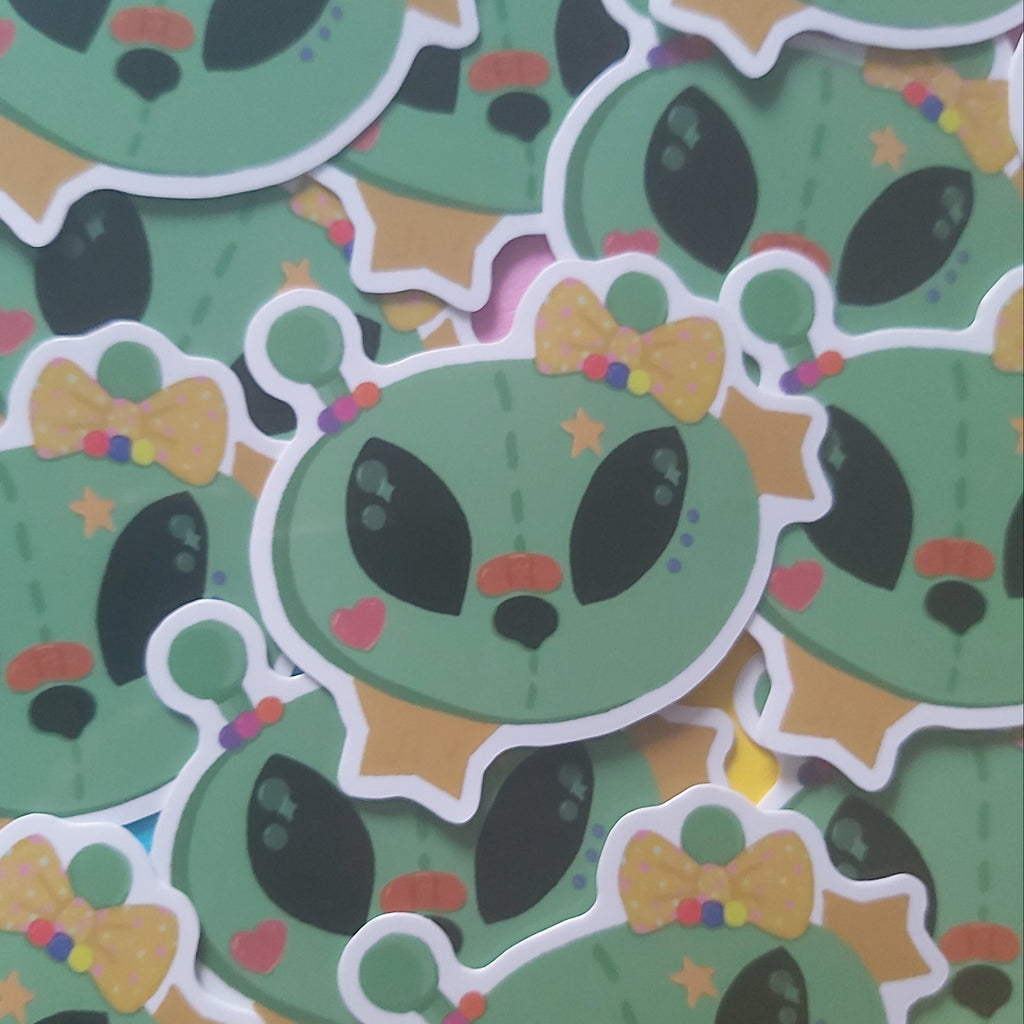 A pile of die-cut stickers featuring a cute green alien head design with black eyes, a yellow polka-dot bow tie, and small colorful accents.