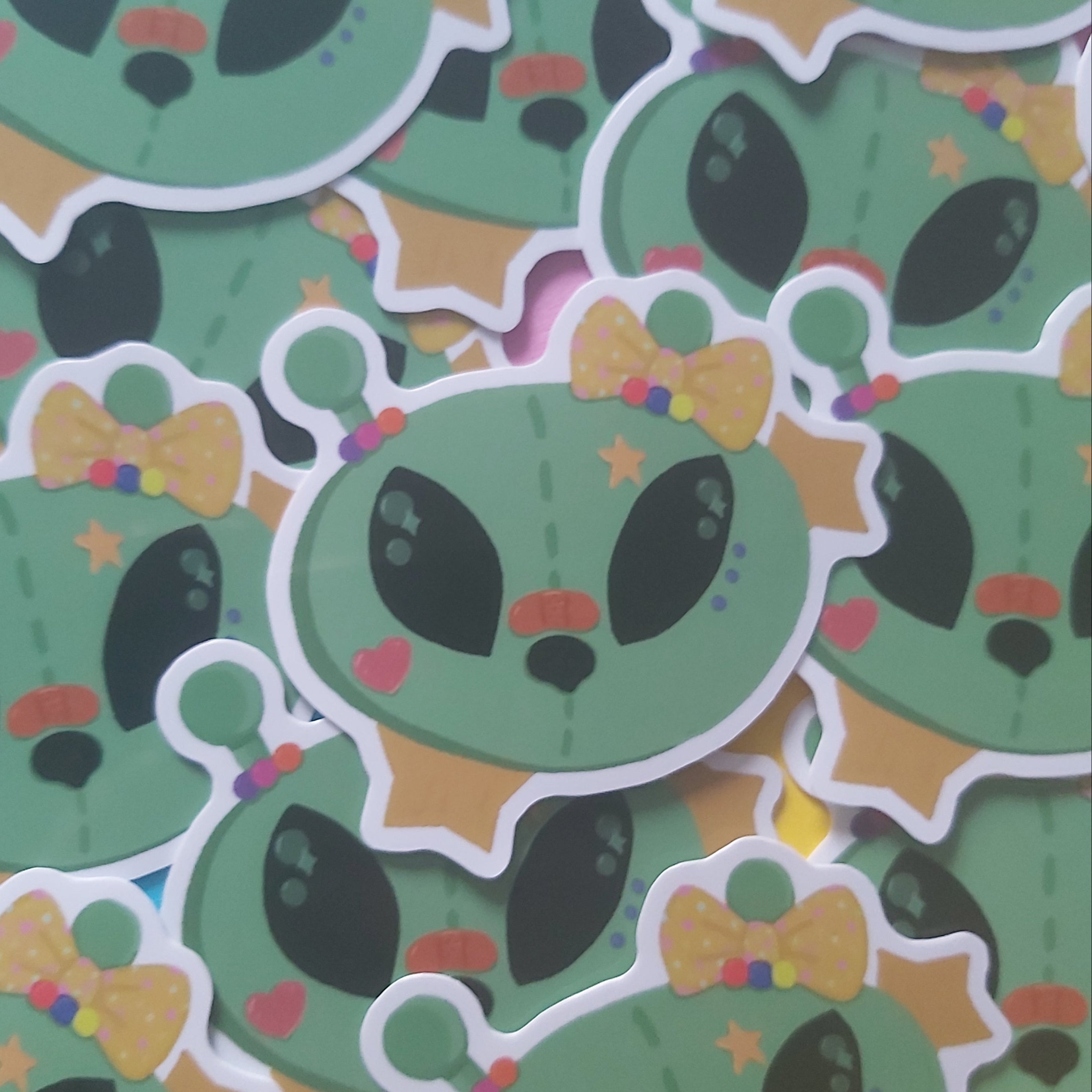 A pile of die-cut stickers featuring a cute green alien head design with black eyes, a yellow polka-dot bow tie, and small colorful accents.
