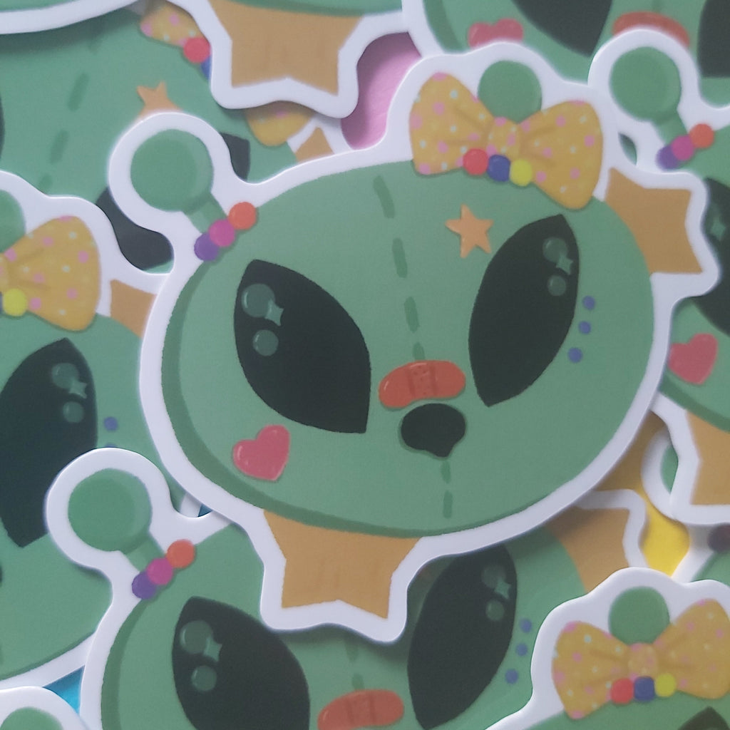 A close-up of multiple cartoon alien stickers layered together. The central sticker shows a green alien face with large black eyes, a small orange nose, pink heart cheeks, a yellow bow tie with multicolored dots, and a small star on its head.