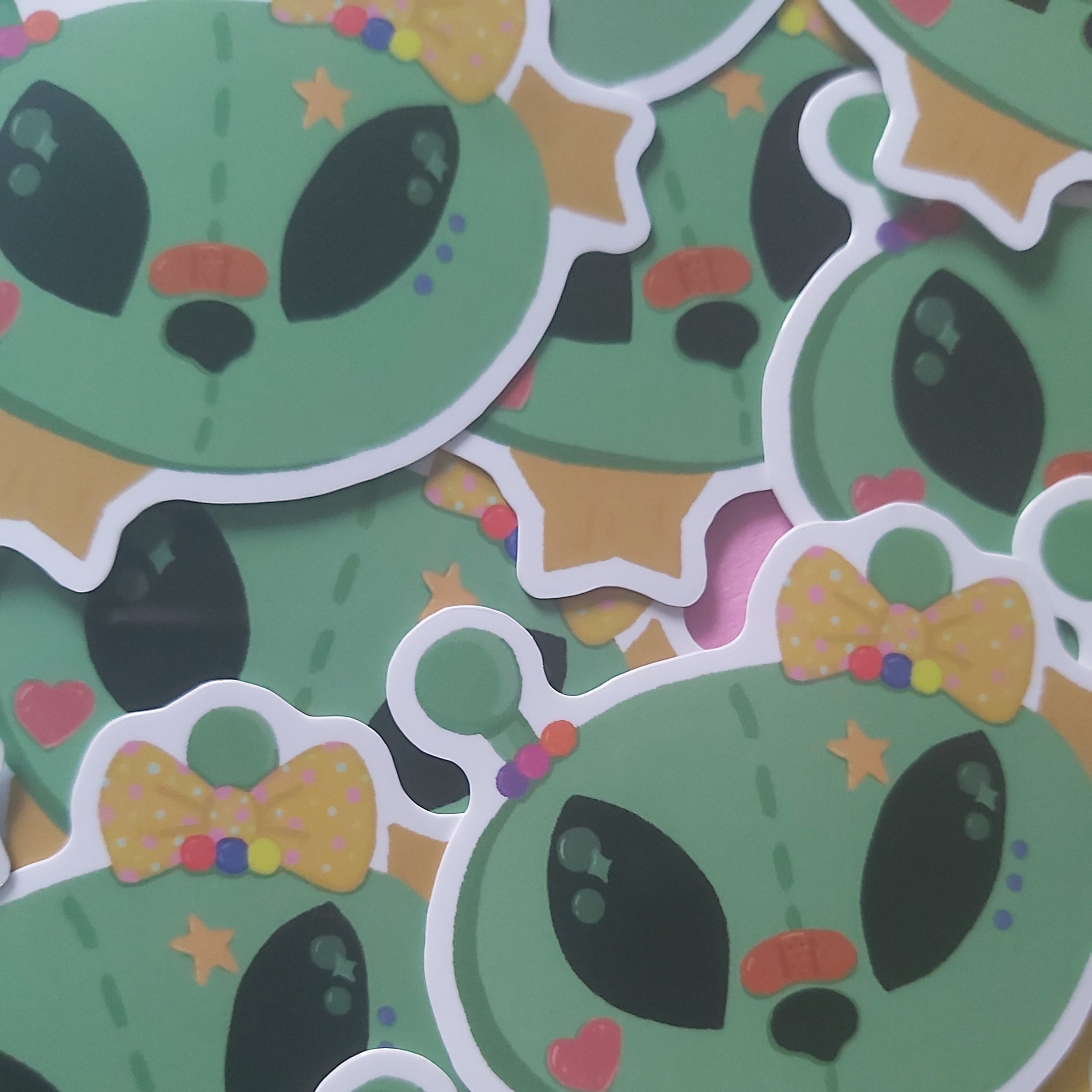 A pile of die-cut stickers featuring a cute green alien design with large black eyes, a yellow polka-dot bow tie, and small colorful accents.