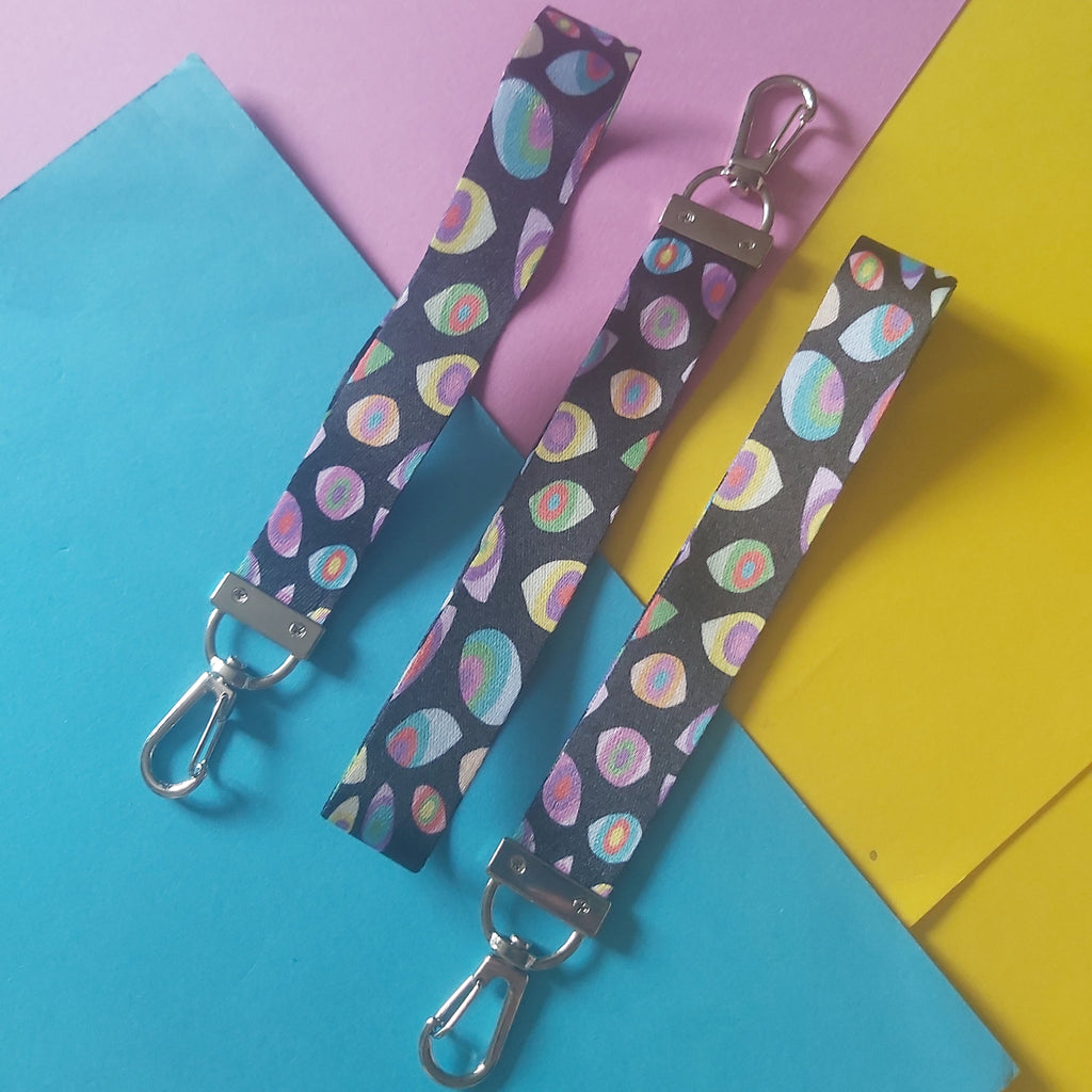 Three fabric wristlet straps with silver metal clasp hardware, featuring a dark background with a colourful pastel eye pattern, arranged on a split pink, blue, and yellow paper background.
