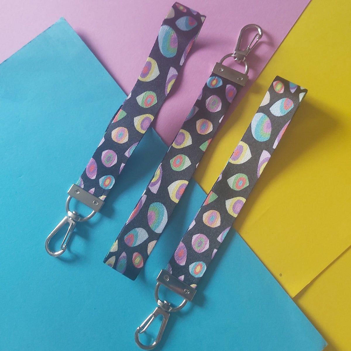 Three fabric wristlet straps with silver metal clasp hardware, featuring a dark background with a colourful pastel eye pattern, arranged on a split pink, blue, and yellow paper background.