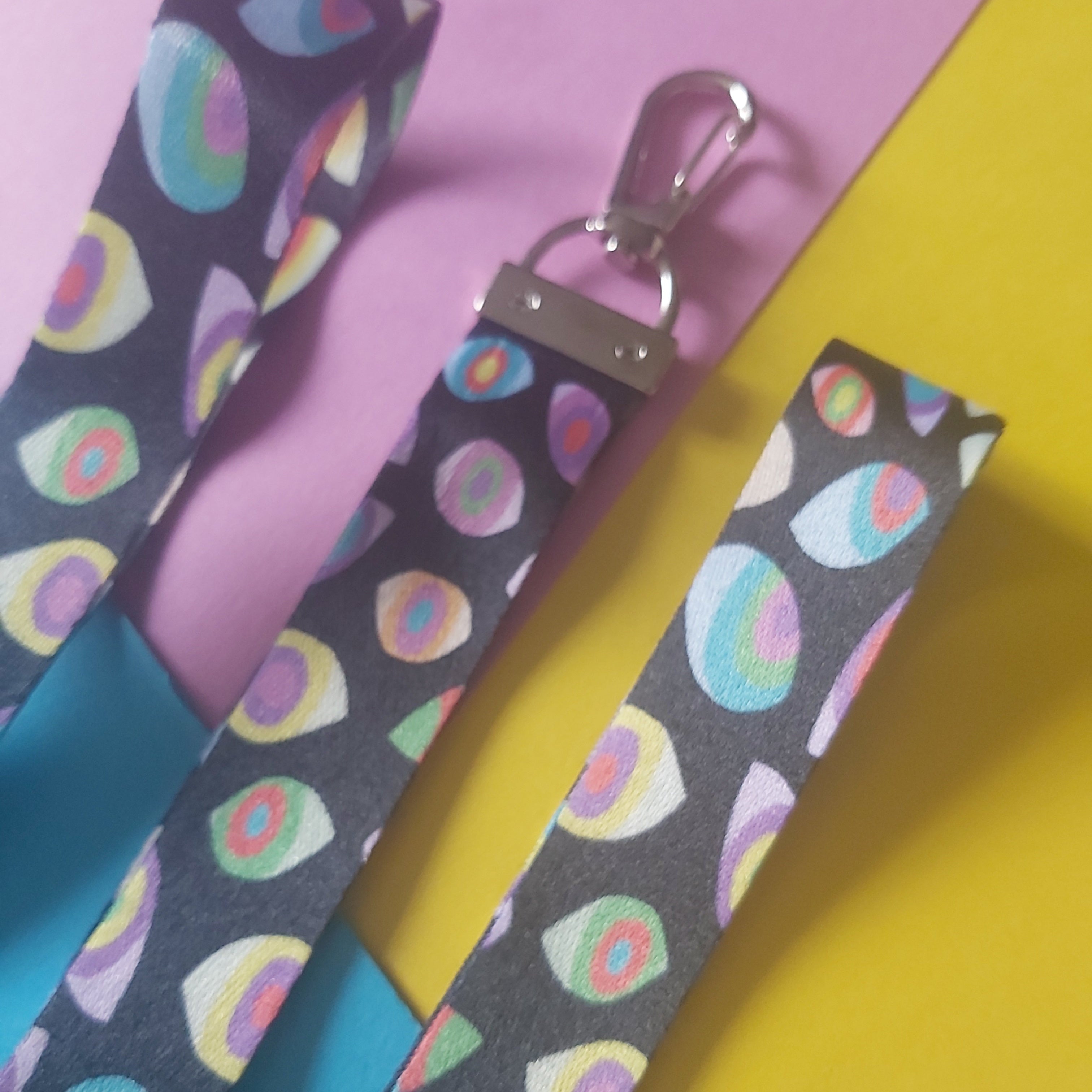 Three fabric dog leashes with a colorful eye pattern on a dark grey background, featuring silver metal clasp hardware, arranged diagonally on a split pink and yellow background.