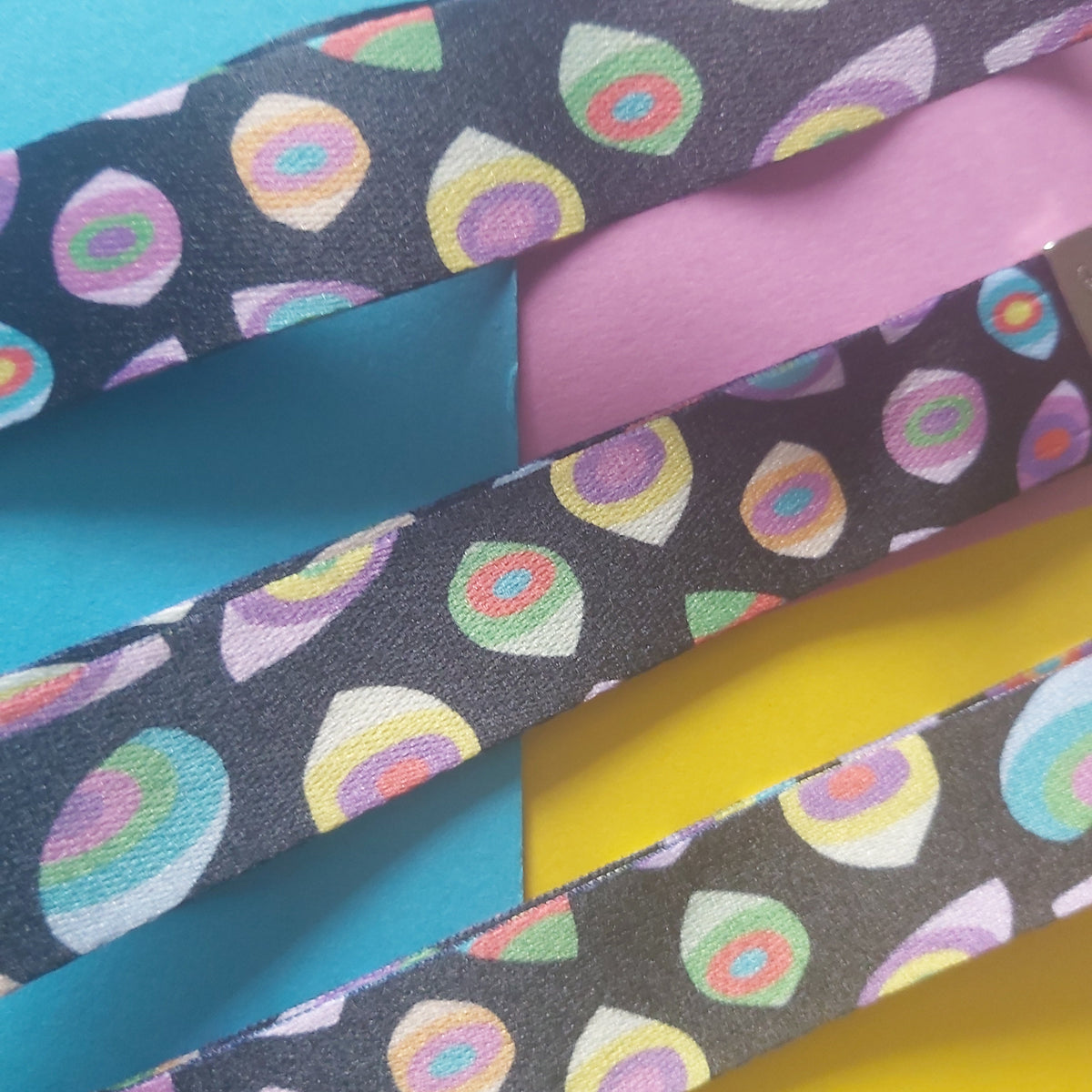 A close-up of two dark grey fabric wristlets with a colourful retro eye pattern, laid diagonally over a background of bright colored paper sheets.