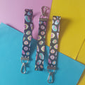 Three fabric wristlet straps with silver metal clips, featuring a colourful pastel rainbow eye pattern on a dark background, arranged on pink, yellow, and blue paper.