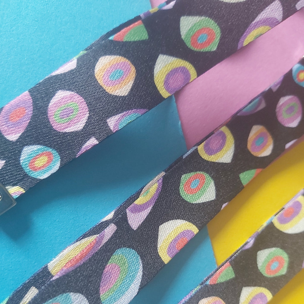 Close-up of three fabric straps laid diagonally on a bright background. The straps are dark charcoal with a repeating multicolour eye pattern in pink, yellow, green, blue, and white.