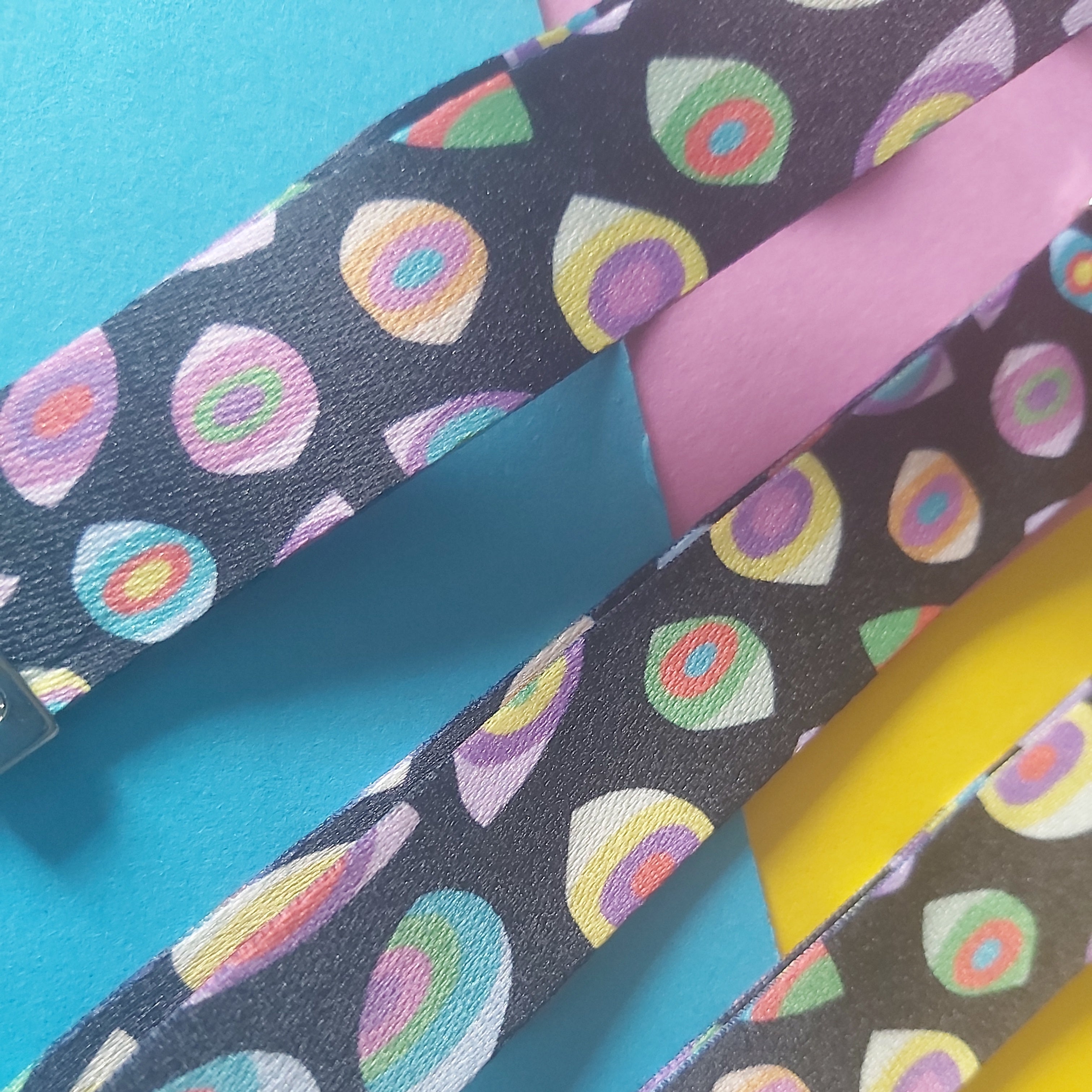 Close-up of three fabric straps laid diagonally on a bright background. The straps are dark charcoal with a repeating multicolour eye pattern in pink, yellow, green, blue, and white.