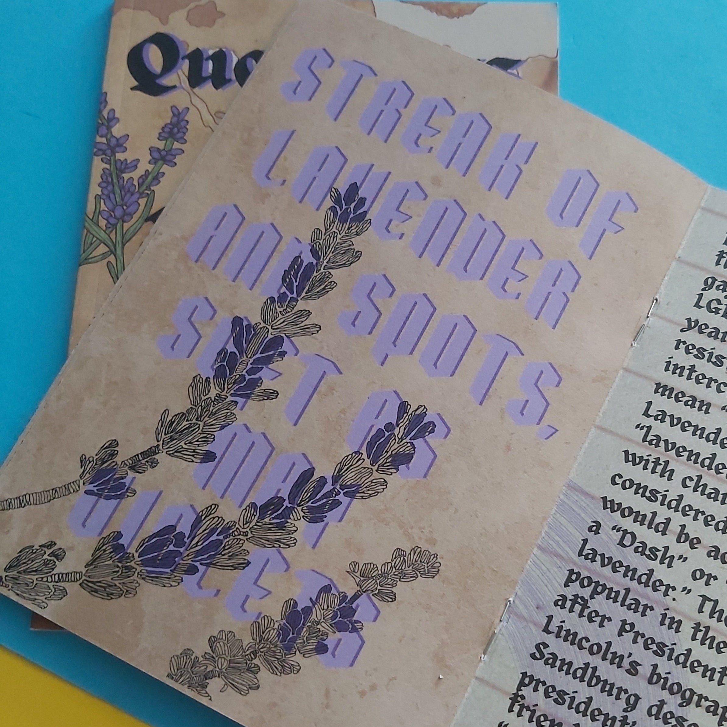 A close-up of zine pages with the phrase “STREAK OF LAVENDER AND SPOTS, SOFT AS MAY VIOLETS” in lavender-purple lettering over a tan background, with lavender illustrations and part of another printed page visible underneath.